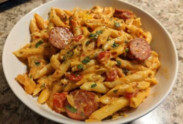 A close-up of a white bowl filled with One-Pot Creamy Cajun Sausage Pasta, featuring penne, creamy sauce, and sliced sausage.