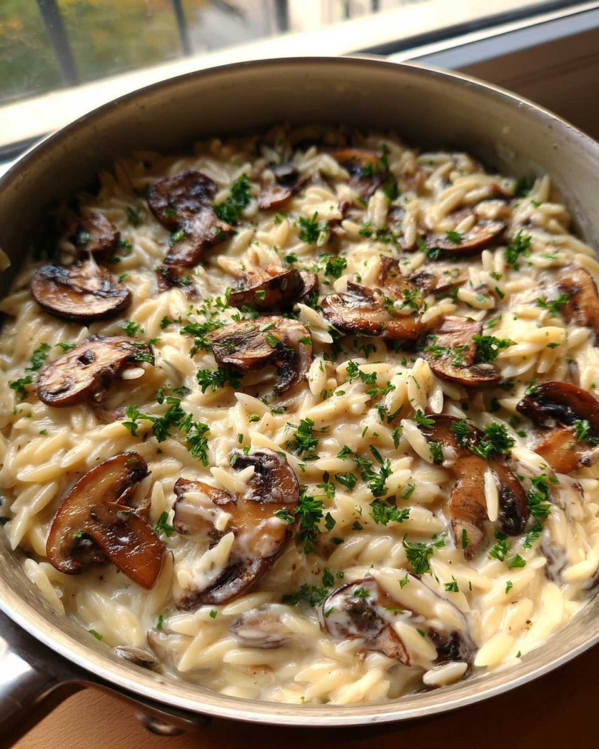 A close-up view of One-Pot Creamy Mushroom Orzo topped with sautéed mushrooms and fresh parsley in a stainless steel pot.