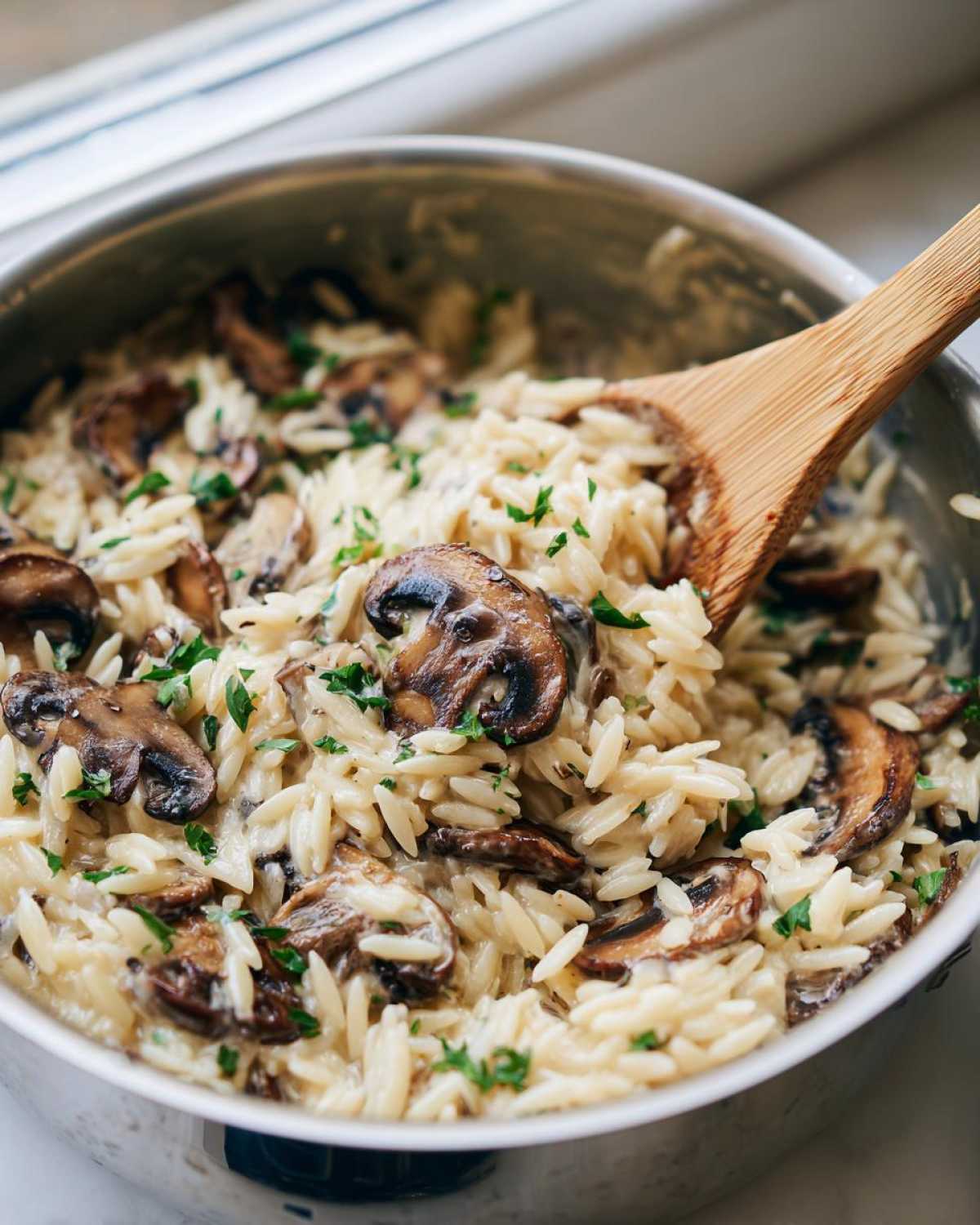 Close-up of creamy orzo pasta mixed with sautéed mushrooms being stirred in a silver pot with a wooden spoon.