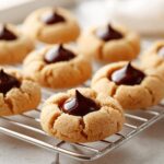 Close-up of freshly baked Peanut Butter Blossom Cookies cooling on a wire rack, topped with melted chocolate kisses.