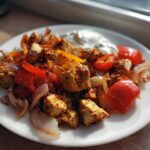 A plate of Sheet Pan Chicken Shawarma mixed with roasted red peppers and onions, served with a dollop of garlic yogurt sauce.