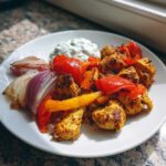A white plate featuring seasoned Sheet Pan Chicken Shawarma pieces mixed with roasted red and yellow peppers and red onion wedges, served with garlic yogurt sauce.