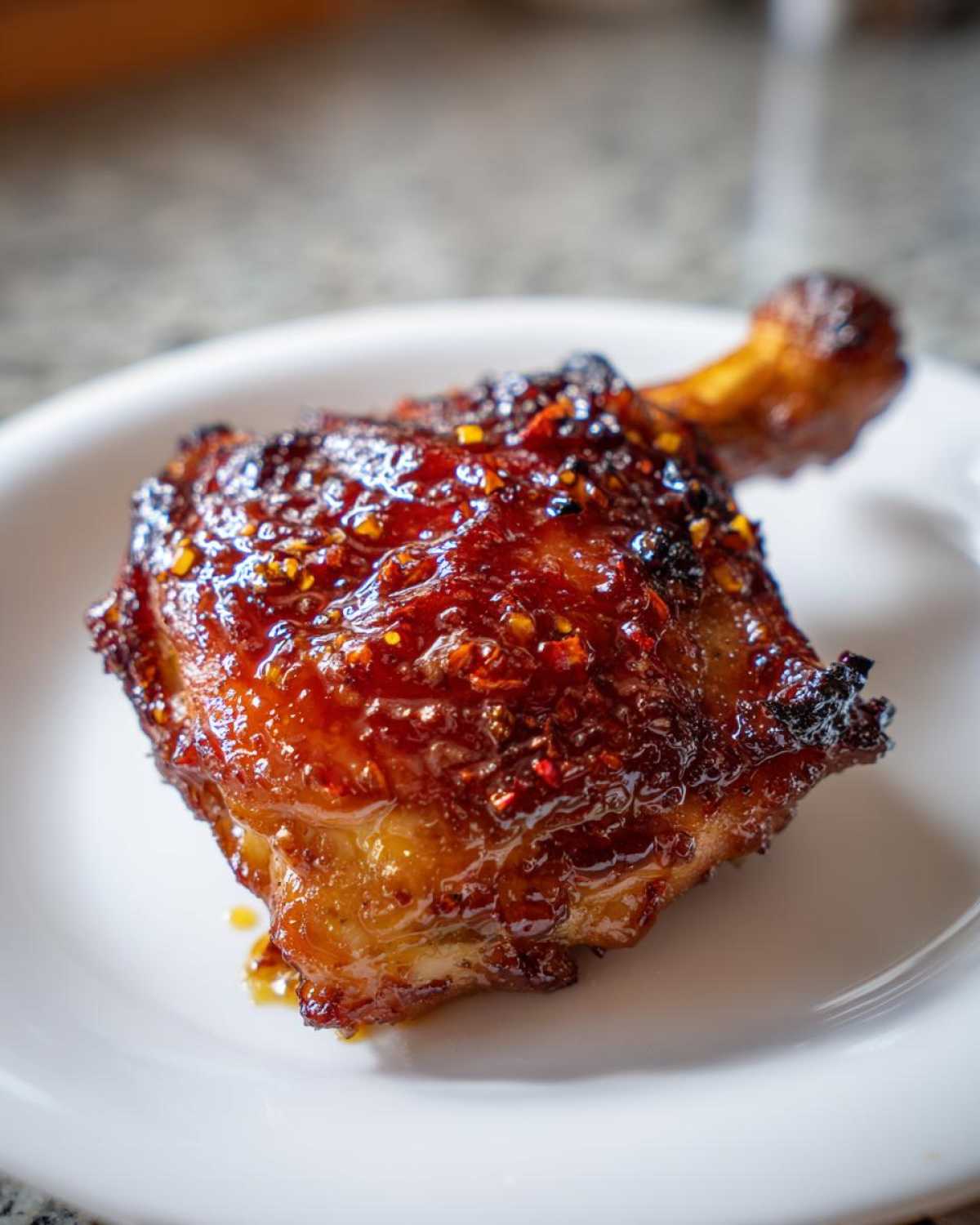 Close-up of a single, sticky, glazed Spicy Honey Garlic Air Fryer Chicken Thigh on a white plate.
