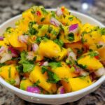 Close-up of a white bowl filled with bright yellow Spicy Mango Salsa, featuring diced mango, red onion, and cilantro.