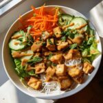 Overhead view of a Spring Roll Bowl with Peanut Sauce, featuring crispy tofu, rice noodles, cucumber, carrots, and mint.