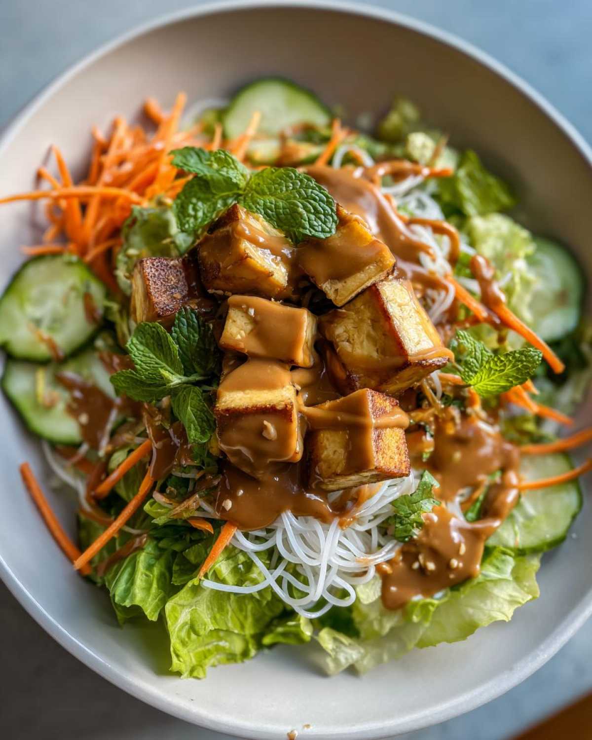 Close-up of a vibrant Spring Roll Bowl with Peanut Sauce, featuring crispy tofu, rice noodles, lettuce, carrots, and mint.