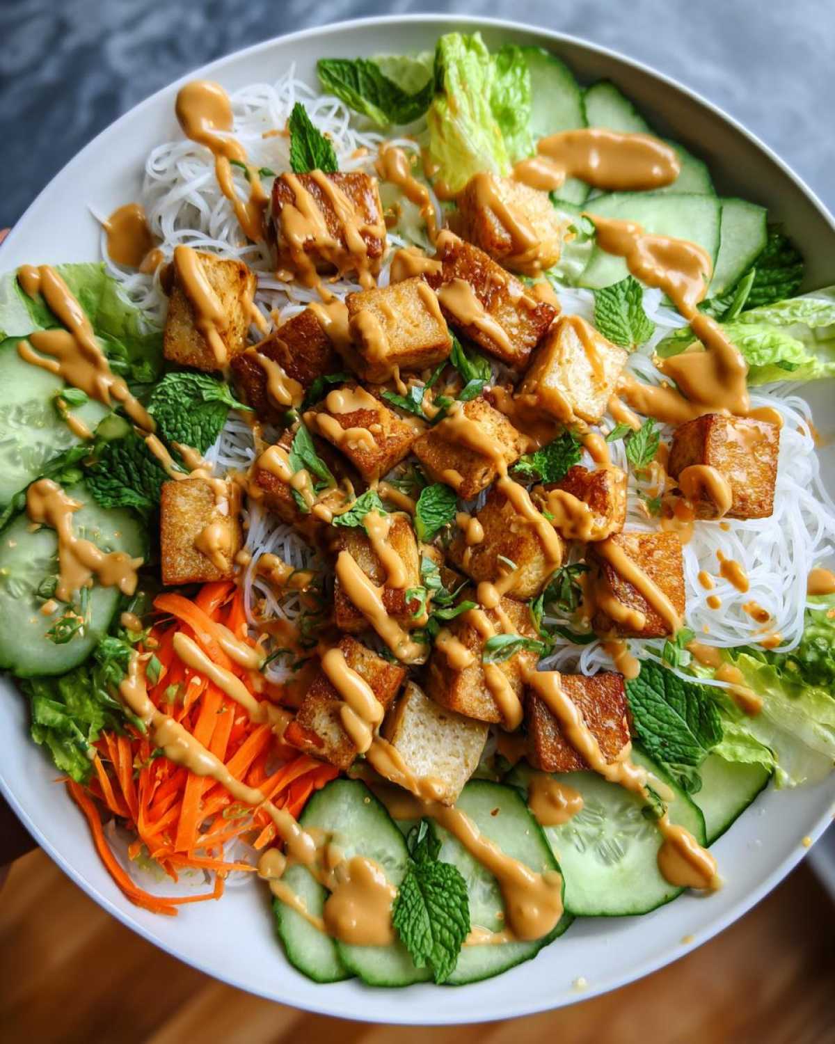 Overhead view of a vibrant Spring Roll Bowl with Peanut Sauce, featuring rice vermicelli, crispy tofu, cucumbers, carrots, and mint.