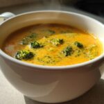 Close-up of a steaming white mug filled with bright orange Copycat Broccoli Cheddar Soup, featuring visible florets of green broccoli.