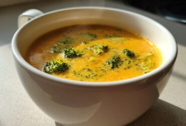 Close-up of a steaming white mug filled with bright orange Copycat Broccoli Cheddar Soup, featuring visible florets of green broccoli.