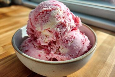 Three scoops of pink and white swirled Strawberry Banana Ice Cream in a speckled bowl.