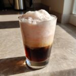 A tall glass showing the layers of a Strawberry Cream Cold Brew with dark coffee at the bottom and thick pink foam on top.