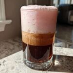 A tall glass showing layered Strawberry Cream Cold Brew with dark coffee, light foam, and thick pink foam on top.