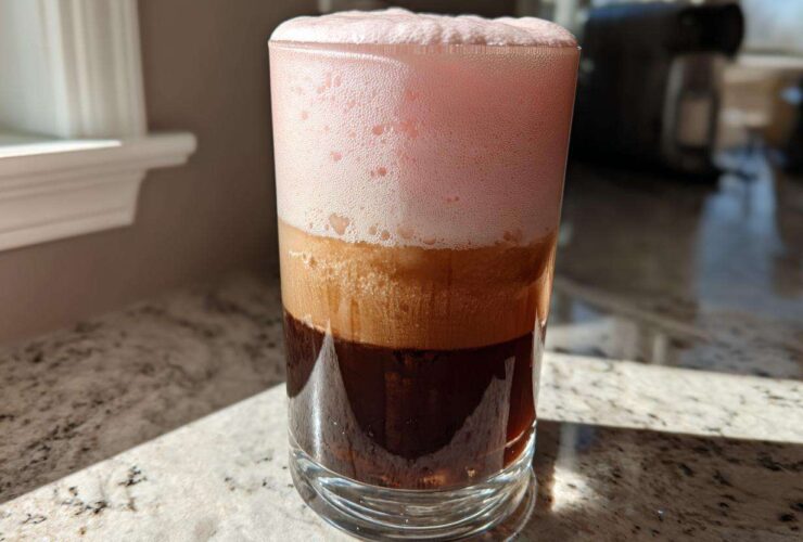 A tall glass showing layered Strawberry Cream Cold Brew with dark coffee, light foam, and thick pink foam on top.