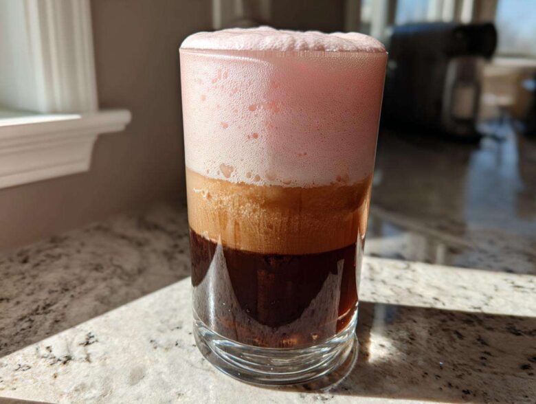 A tall glass showing layered Strawberry Cream Cold Brew with dark coffee, light foam, and thick pink foam on top.
