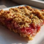 A close-up, sunlit view of a square slice of Strawberry Rhubarb Crisp showing the thick, gooey fruit filling and crunchy oat topping.