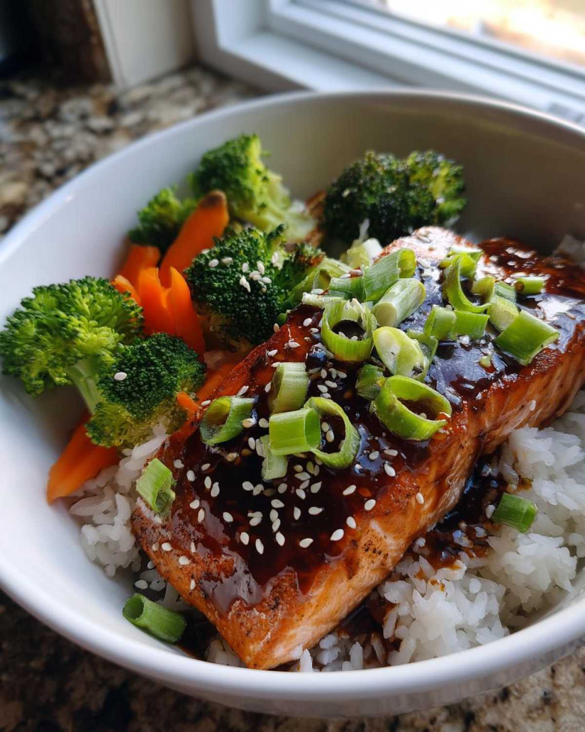 A close-up of a Teriyaki Salmon Rice Bowls serving, featuring glazed salmon over rice with broccoli, carrots, and green onions.