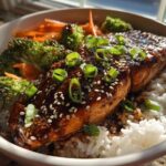 A close-up of a Teriyaki Salmon Rice Bowls featuring a glazed salmon fillet over white rice with broccoli and carrots.