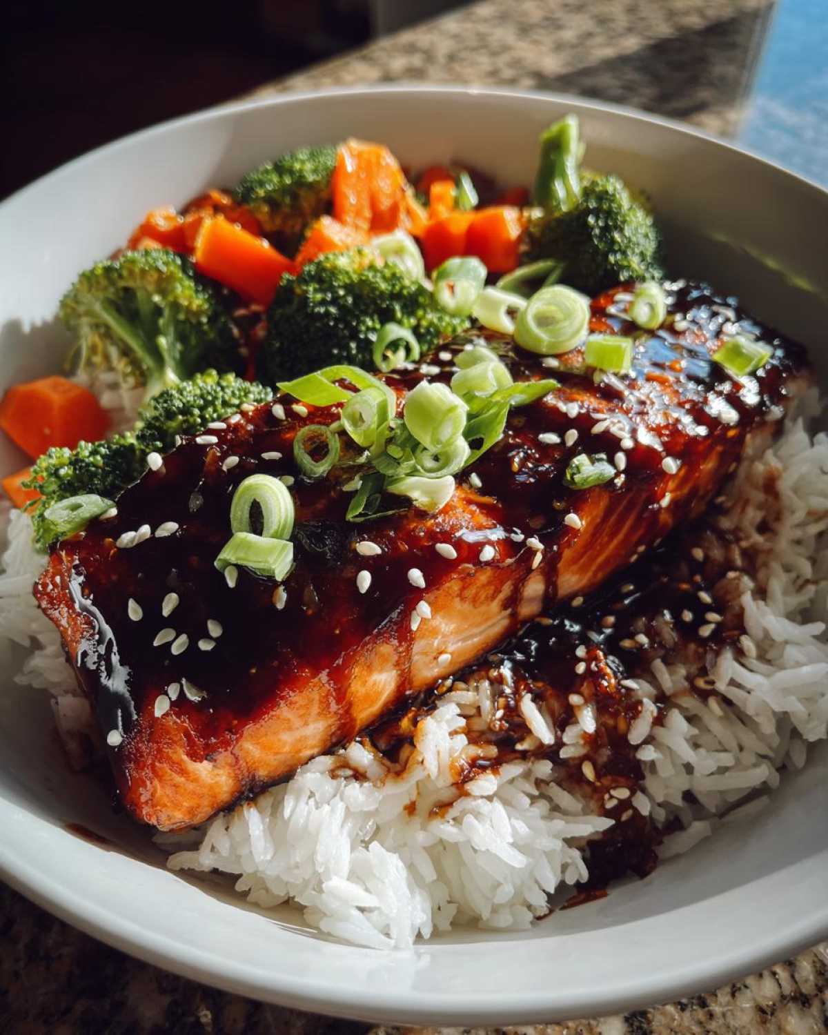 A close-up of a Teriyaki Salmon Rice Bowls serving featuring glazed salmon over white rice with broccoli and carrots.
