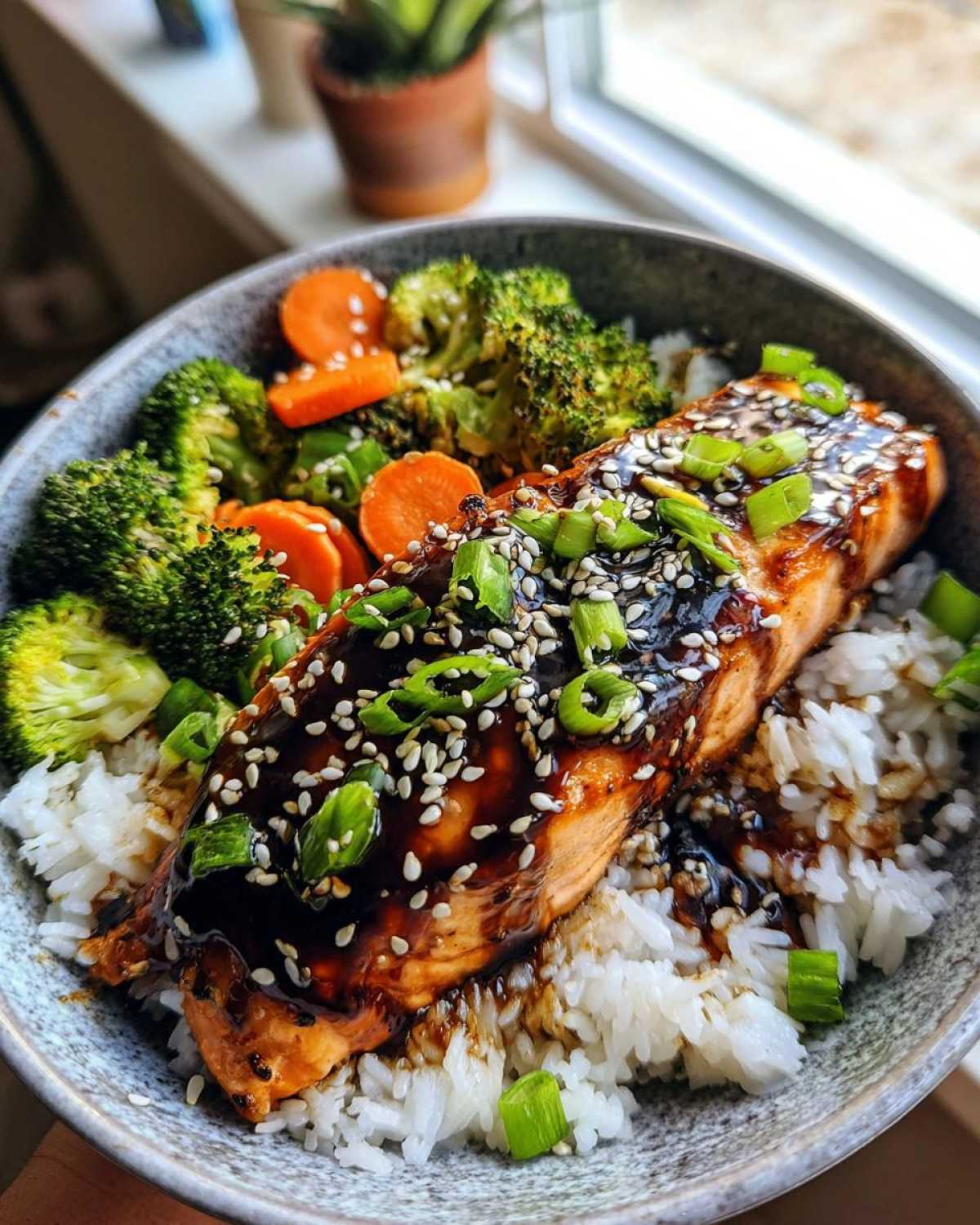 A close-up of a Teriyaki Salmon Rice Bowls serving featuring glazed salmon over white rice with broccoli, carrots, and sesame seeds.