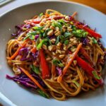 A close-up of a bowl of Thai Peanut Noodles with Crunchy Veggies, topped with peanuts and scallions.