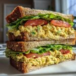 A tall, stacked Vegan Chickpea Salad Sandwich featuring yellow chickpea filling, lettuce, and tomato on whole grain bread.