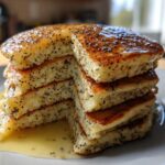 A tall stack of fluffy Vegan Lemon Poppy Seed Pancakes drizzled with lemon syrup.