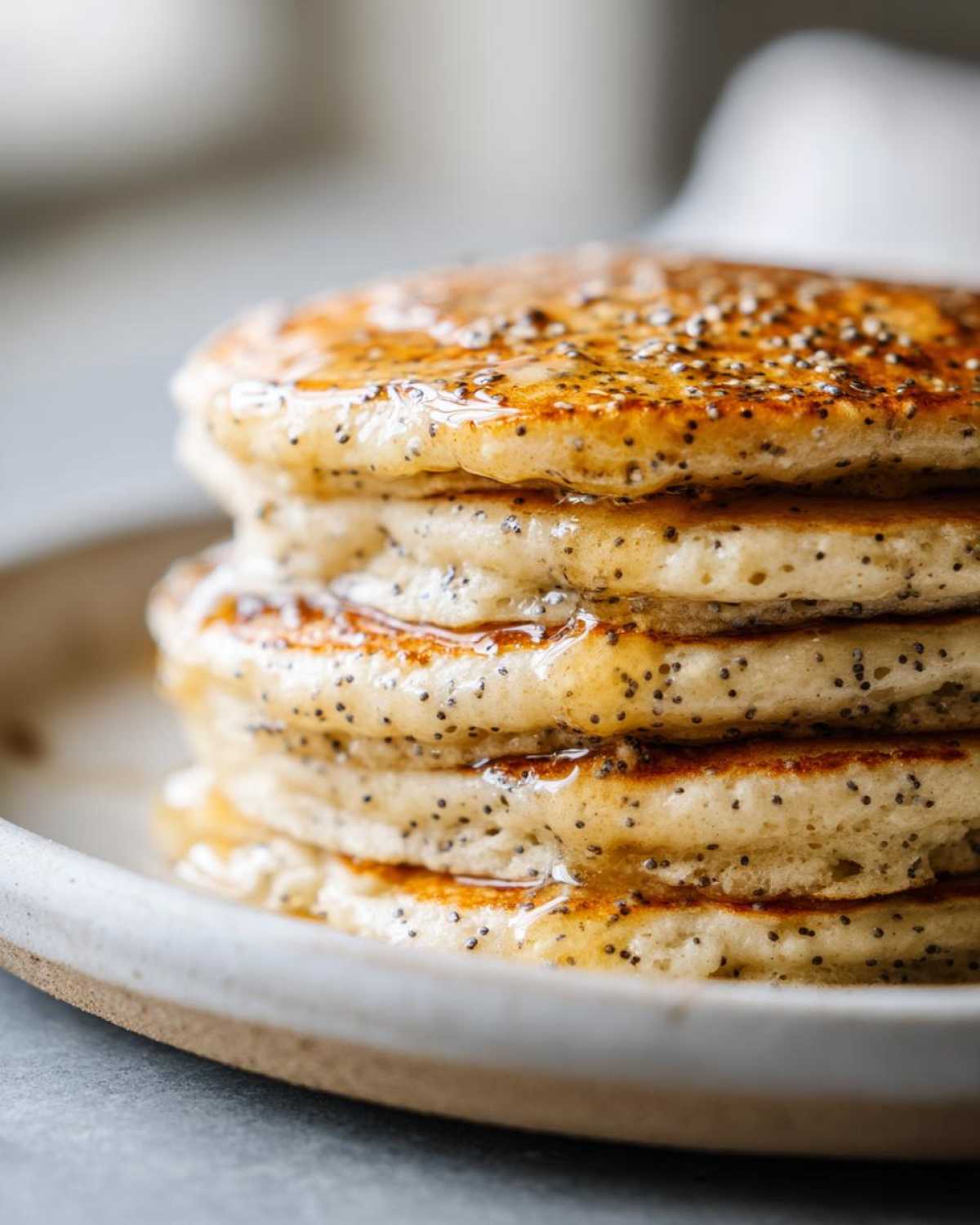 Close-up of a tall stack of fluffy Vegan Lemon Poppy Seed Pancakes drizzled with syrup and speckled with poppy seeds.