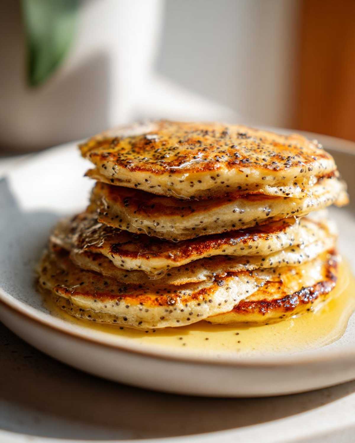 A tall stack of golden Vegan Lemon Poppy Seed Pancakes drizzled generously with syrup.
