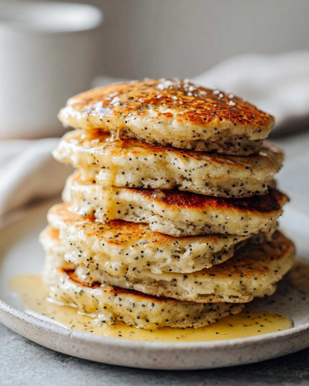 Close-up of a tall stack of fluffy Vegan Lemon Poppy Seed Pancakes drizzled with syrup.