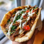 Close-up of a handheld Vegan Mushroom Shawarma stuffed with shredded mushrooms, tomatoes, parsley, and drizzled with white sauce.