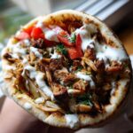 Close-up of a handheld Vegan Mushroom Shawarma stuffed with seasoned mushrooms, tomatoes, and white sauce.