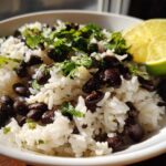 Close-up of a bowl of fluffy white rice mixed with black beans, topped with fresh cilantro and a lime wedge, showcasing the beans and rice dish.