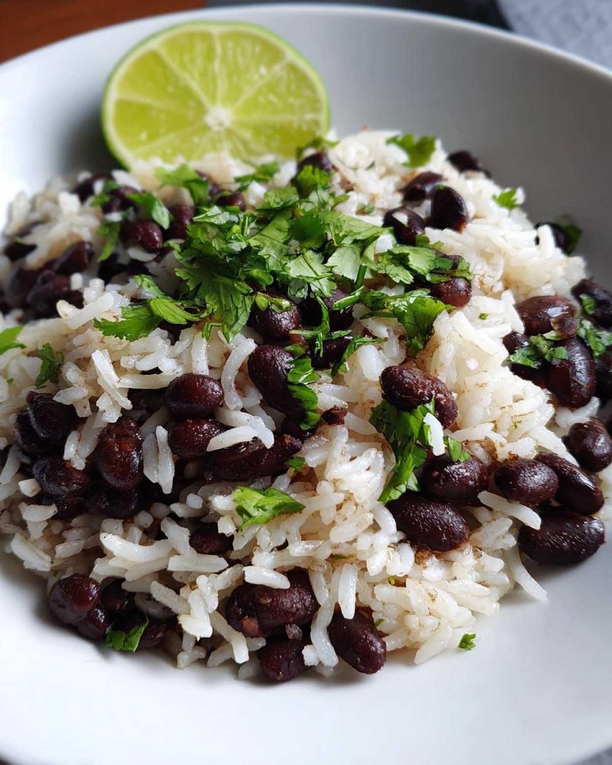 Close-up of a bowl of fluffy white rice mixed with black beans, topped with fresh cilantro and a lime wedge for the perfect beans and rice.