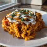A serving of cheesy taco pasta featuring rotini noodles, ground meat sauce, melted cheddar, sour cream, and fresh parsley.