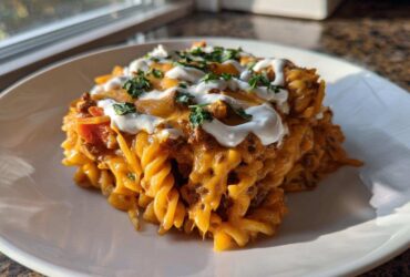 A serving of cheesy taco pasta featuring rotini noodles, ground meat sauce, melted cheddar, sour cream, and fresh parsley.