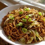 A close-up of a white bowl filled with glossy, brown fried noodles, mixed with cabbage and topped with bright green scallions.