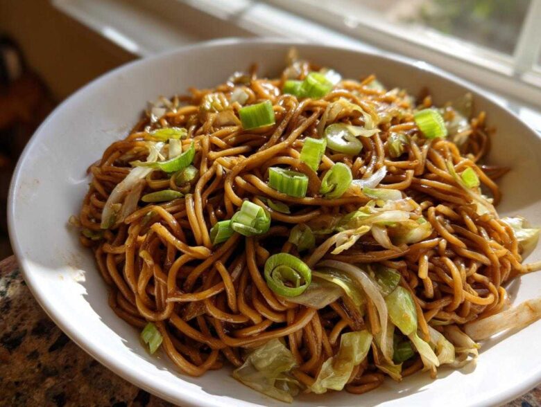 A close-up of a white bowl filled with glossy, brown fried noodles, mixed with cabbage and topped with bright green scallions.