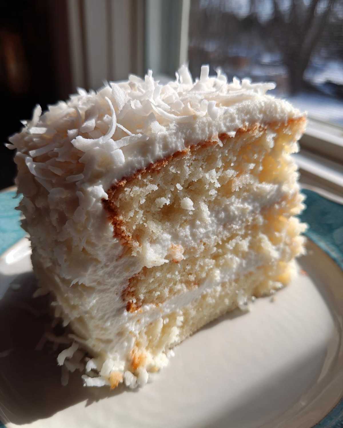 Close-up of a moist slice of 3-layer coconut cake recipe, heavily frosted and topped with shredded coconut.