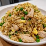 A close-up, appetizing shot of a mound of homemade chicken fried rice with visible pieces of chicken, green peas, and scrambled egg.