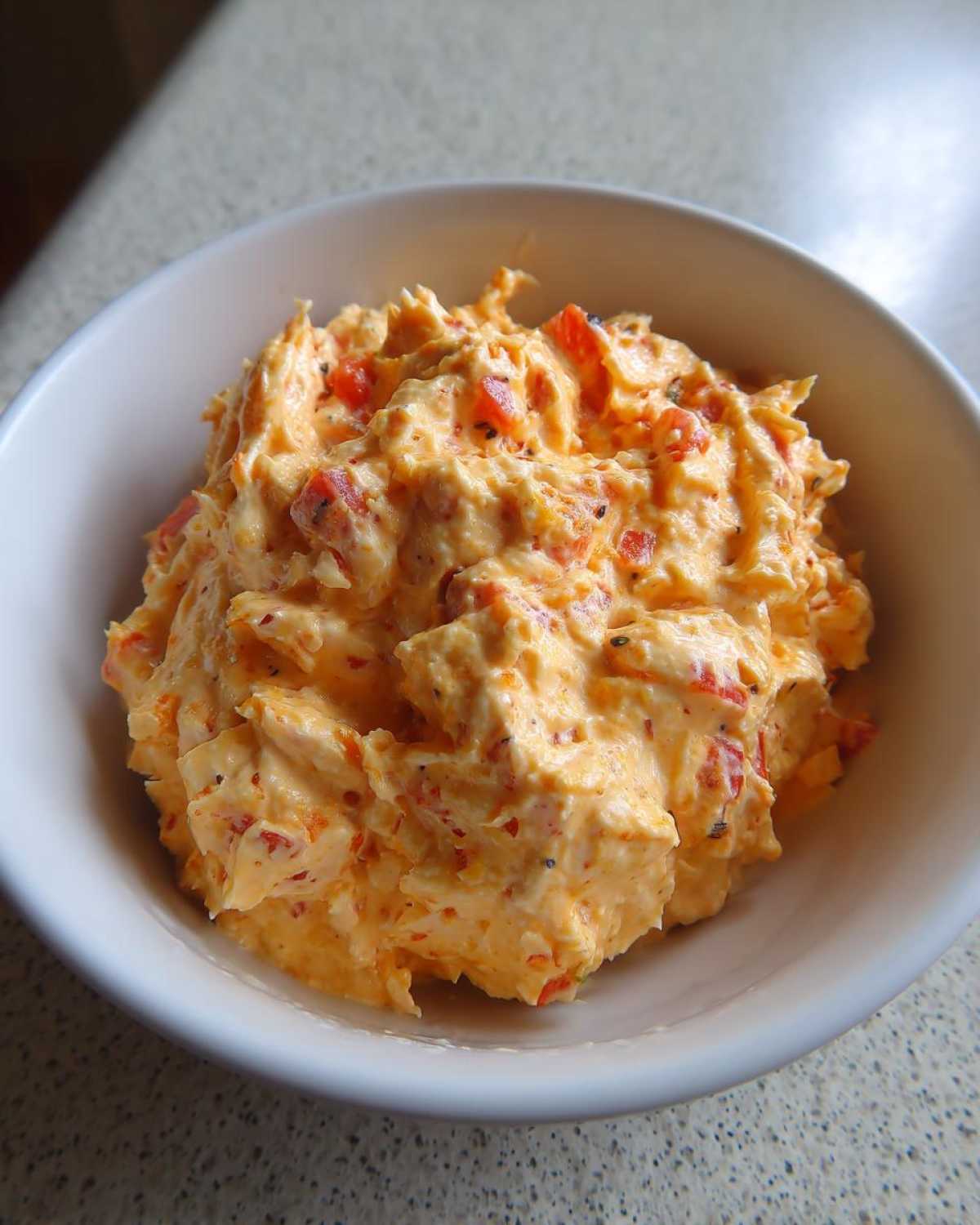 A close-up of thick, orange-colored boat dip mixed with red peppers served in a small white bowl.