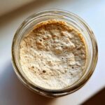 Overhead view of an active, bubbly sourdough starter inside a clear glass jar, showing signs of fermentation.