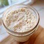 Close-up overhead view of an active, bubbly sourdough starter in a glass jar, showing visible oat flakes.