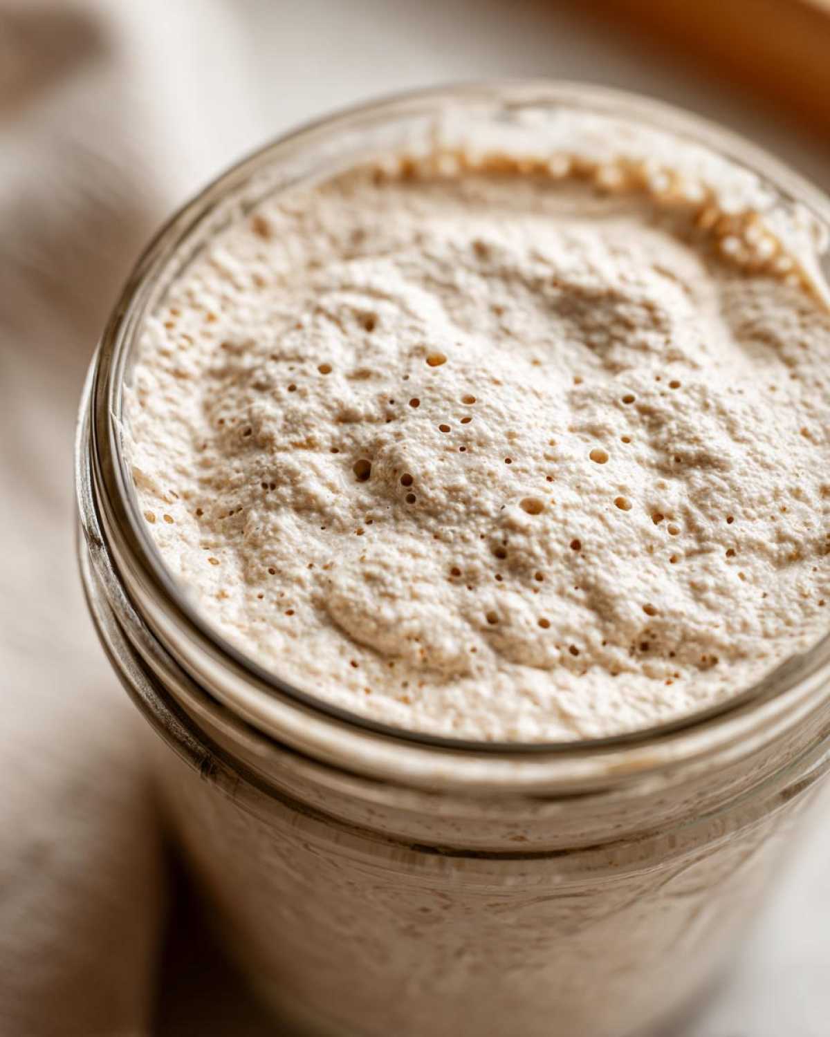 Close-up view of an active, bubbly sourdough starter overflowing slightly from the top of a clear glass jar.