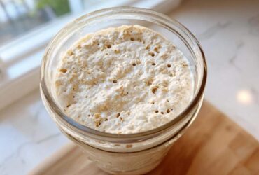 Close-up overhead view of an active, bubbly sourdough starter in a glass jar, showing visible oat flakes.