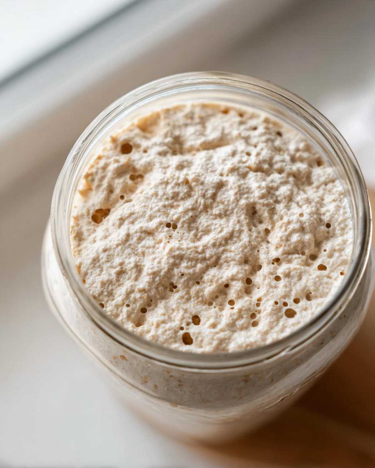 Overhead view of an active sourdough starter, showing a bubbly, risen texture inside a clear glass jar.