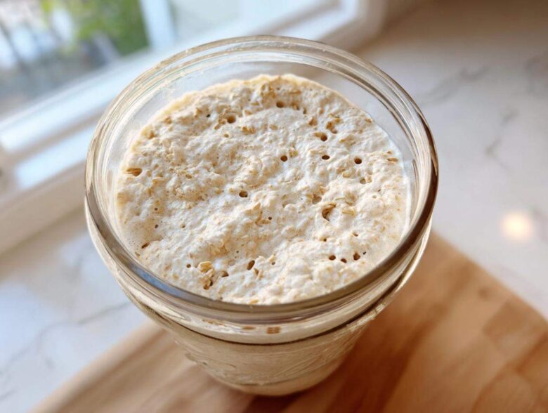 Close-up overhead view of an active, bubbly sourdough starter in a glass jar, showing visible oat flakes.