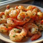 A close-up of cooked, seasoned air fryer frozen shrimp piled on a light-colored plate, glistening with butter and herbs.