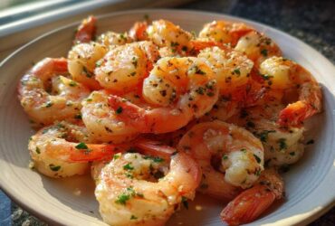 A close-up of cooked, seasoned air fryer frozen shrimp piled on a light-colored plate, glistening with butter and herbs.