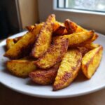 A close-up of a pile of crispy, golden-brown air fryer potato wedges seasoned with spices, served on a white plate.