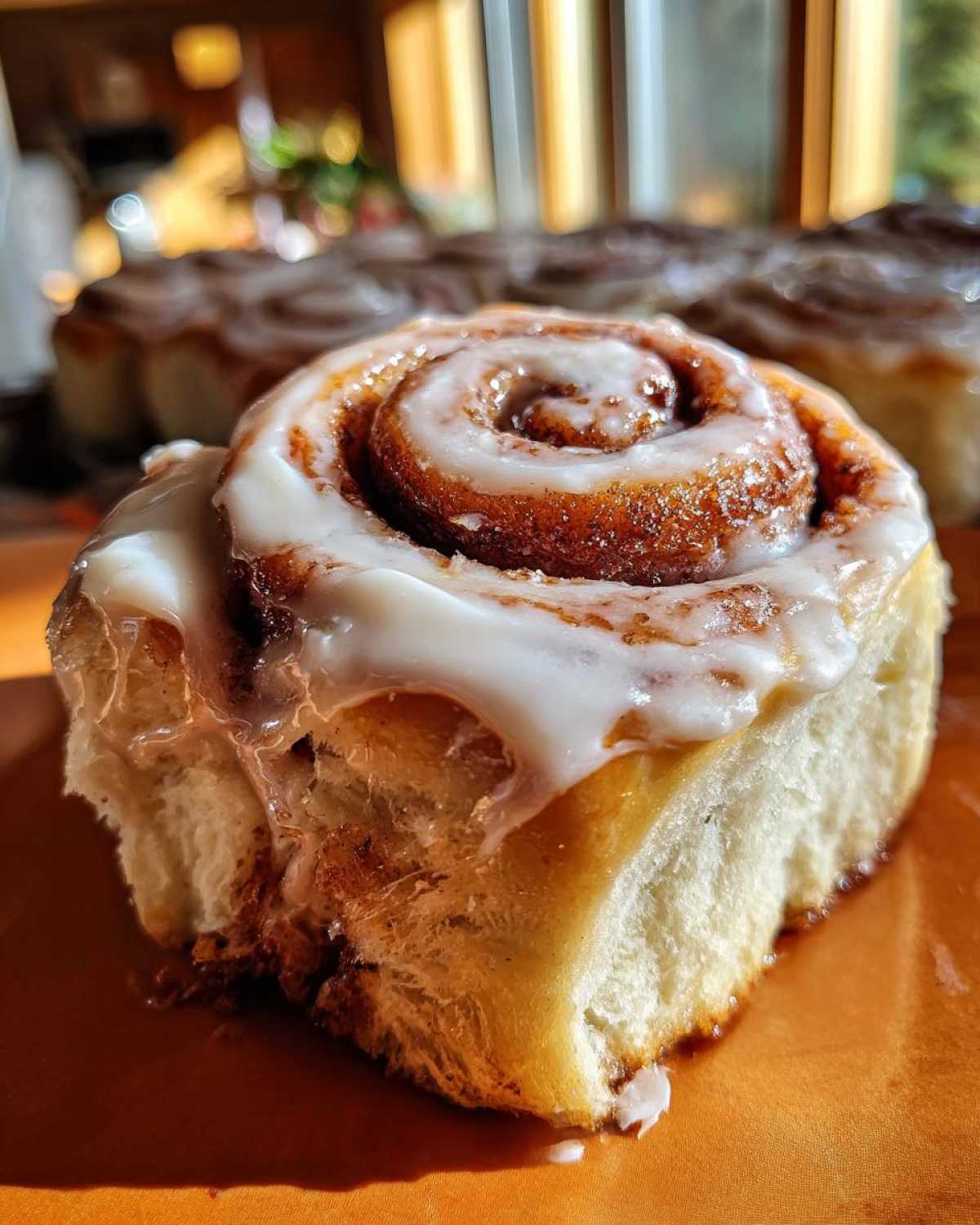 A close-up of a soft, freshly baked cinnamon bun topped with thick white icing, with more cinnamon buns blurred in the background.
